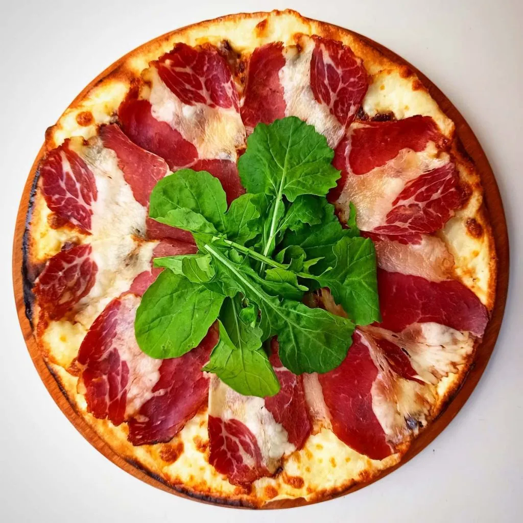 Overhead view of a 400 Derece white-based pizza on a round wooden board, layered with marbled cured meat around a pile of arugula