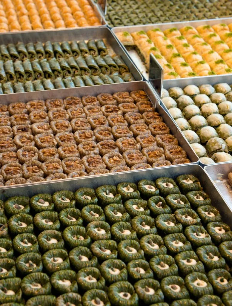 Shop trays of baklava varieties side by side: pistachio rolls, walnut-topped rounds, and cream-filled nests