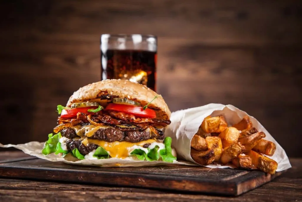 Double cheeseburger with bacon, lettuce, and tomato beside a paper cone of fries and a glass of cola