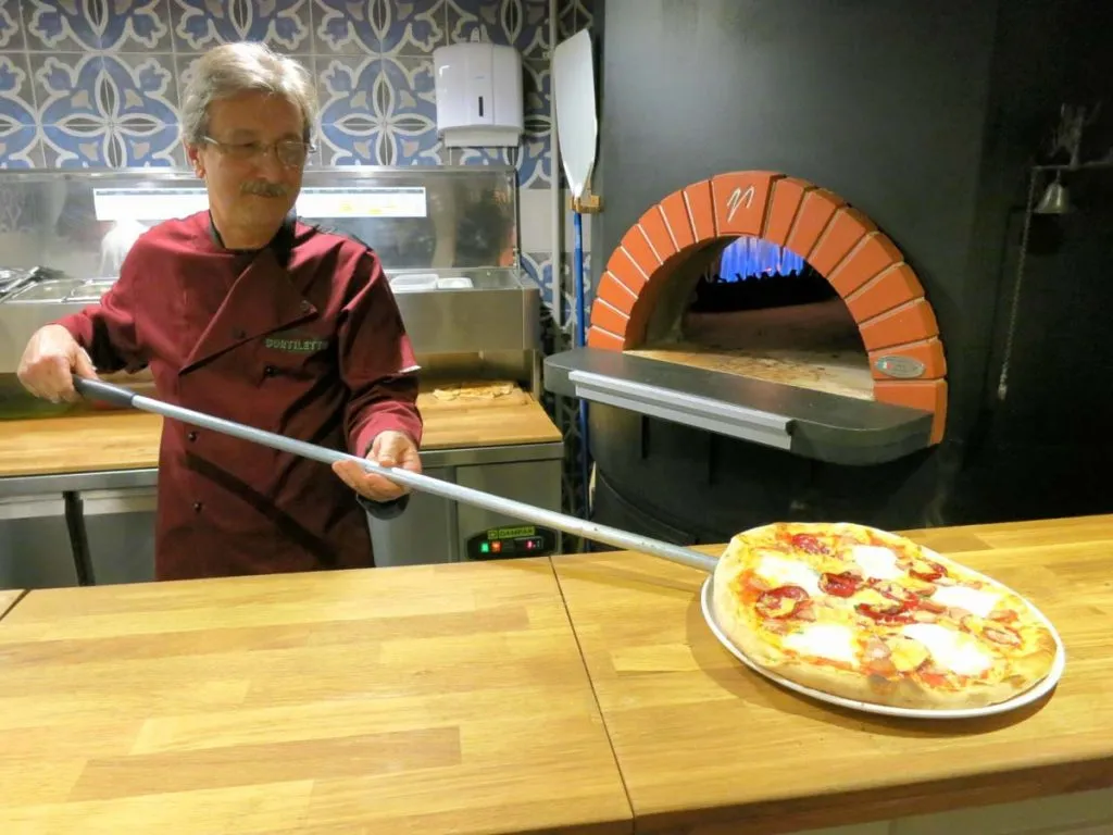 Cortiletto's pizzaiolo pulling a fresh mozzarella pizza out of the brick-arched oven on a long peel