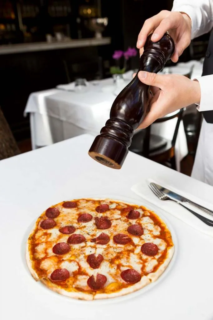 A server grinds black pepper from a tall wooden mill over a salami-topped pizza on a white tablecloth at Da Mario