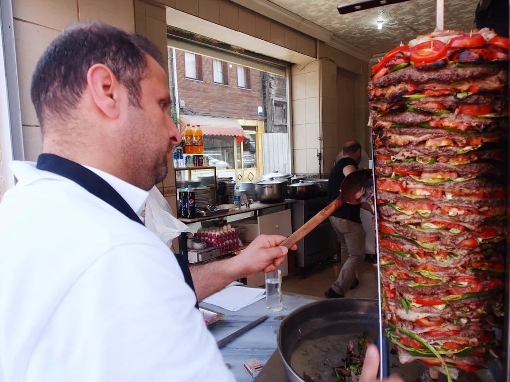 Large roll of beef doner kebab layered with vegetables on rotisserie being shaved by chef