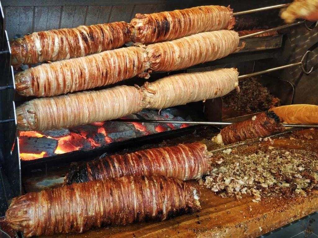 Kokoreç rolls roasting on spits over glowing coals, with chopped kokoreç piled on the cutting board