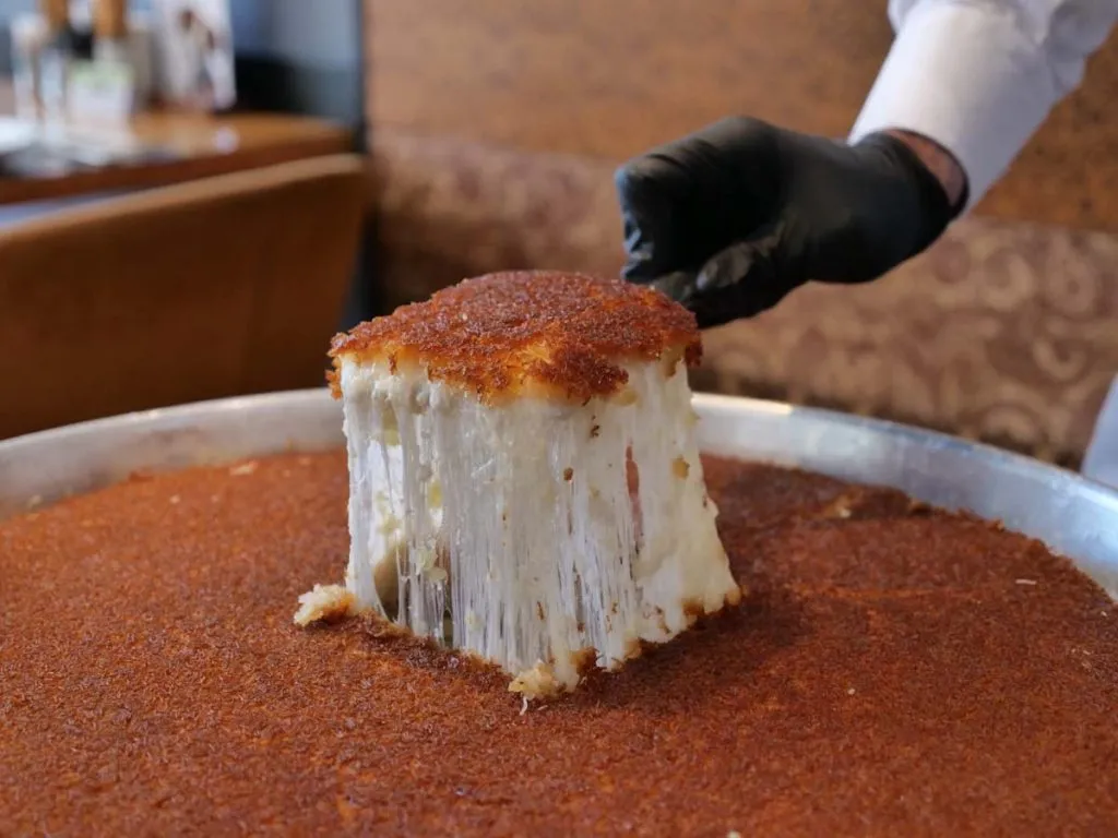Gloved hand lifting a künefe square from a wide tray at Hatay Gurme, the cheese stretching in long strands