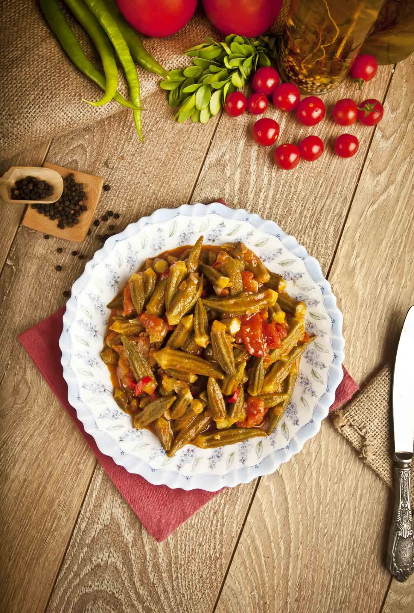 Cooked okra with tomatoes on a patterned white plate, with green peppers and cherry tomatoes nearby