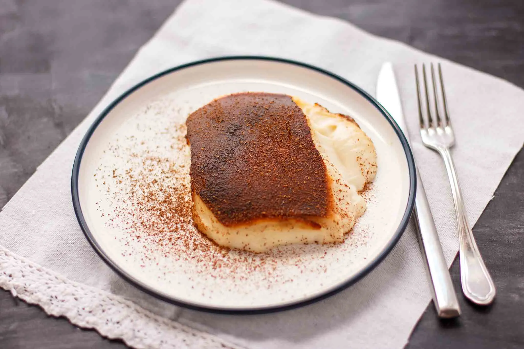 Slice of caramel-topped milk pudding dusted with cinnamon on a plate with fork and knife