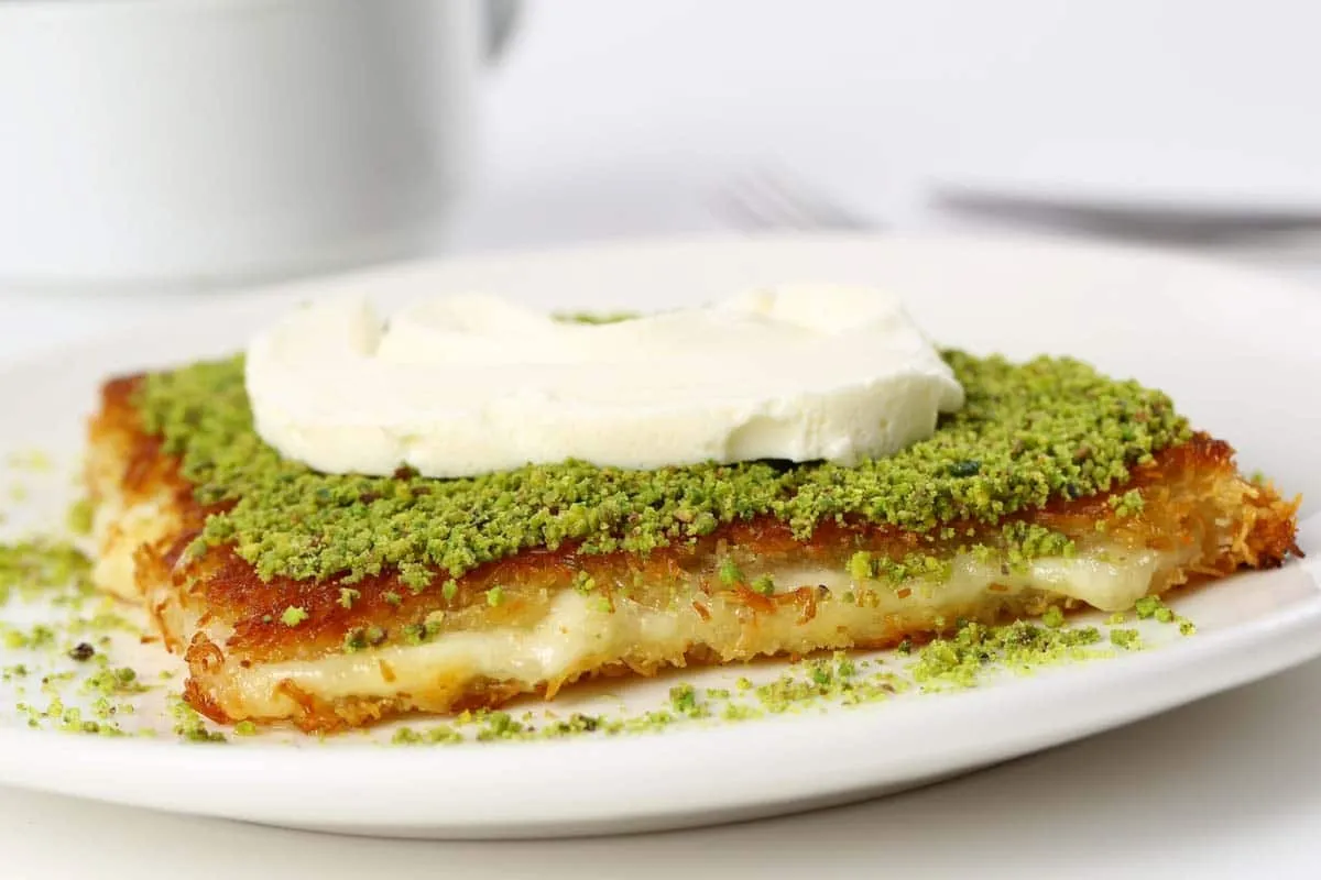 Slice of kunefe topped with ground pistachio and clotted cream, melted cheese inside, on a white plate