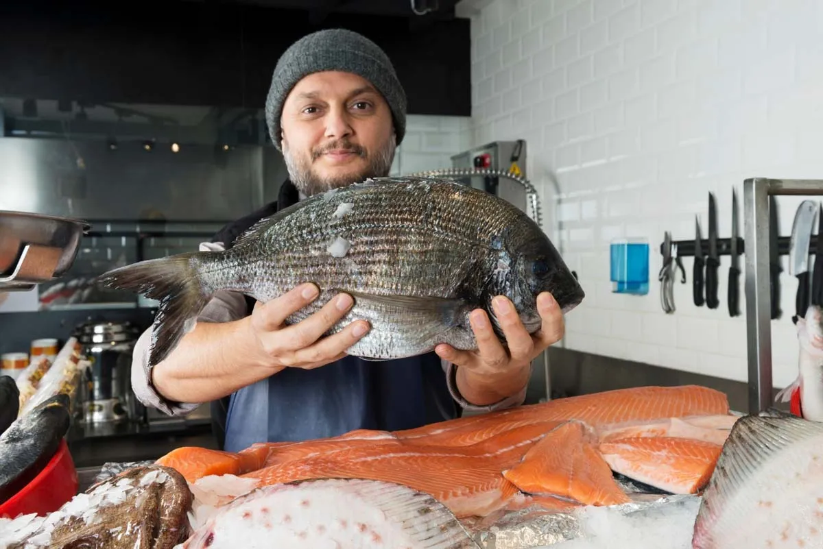 Fishmonger in a gray beanie holding a whole sea bream above salmon fillets and fresh fish