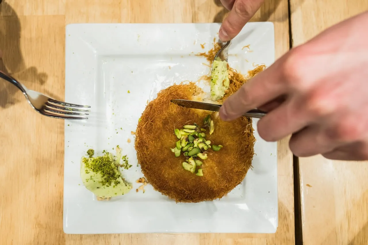 White plate of crisp künefe topped with pistachios being cut with a knife and fork