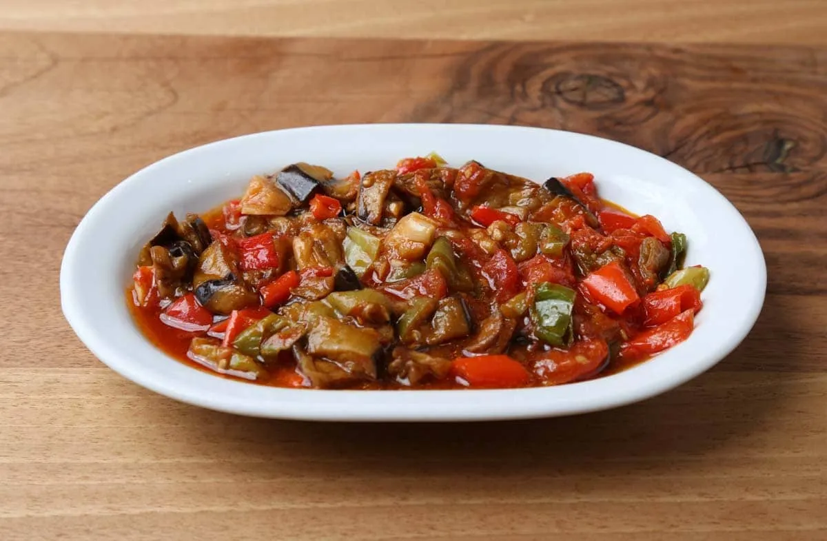 Saksuka with fried eggplant, red and green peppers, and tomato sauce on a white oval plate