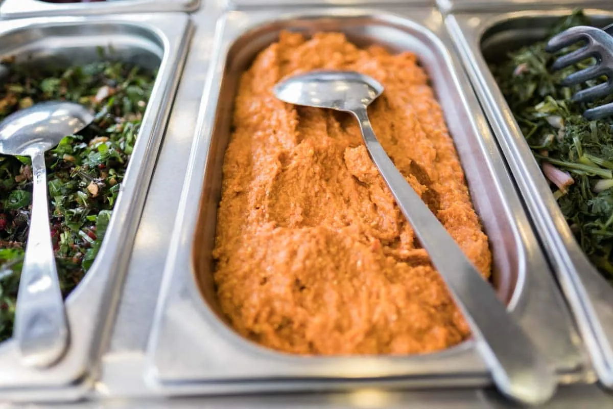 Red muhammara spread in a metal serving tray between two green herb salads with serving spoons