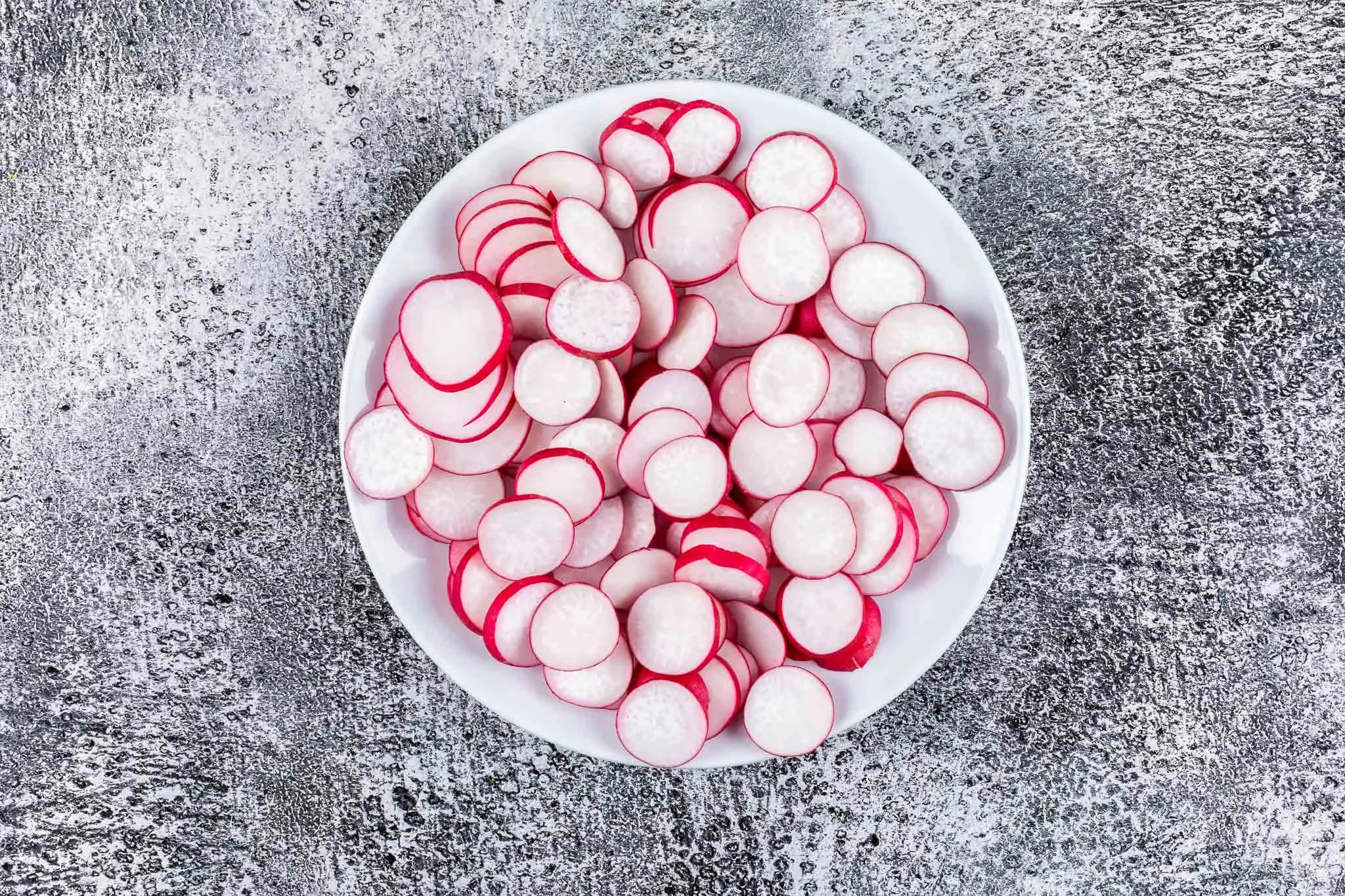 White bowl of thinly sliced red radishes on a gray surface, seen from above