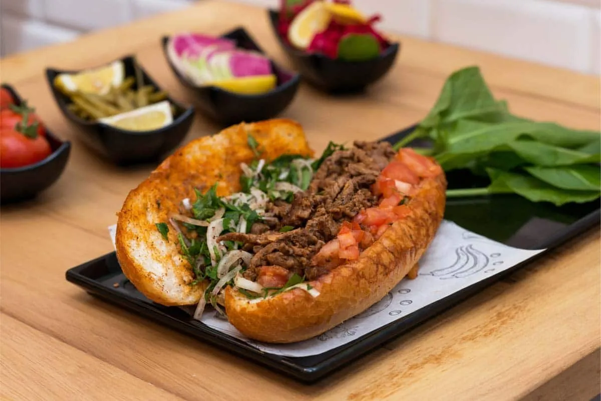 Tantuni served in half a loaf of bread with chopped beef, tomato, onion, and parsley, with arugula and pickled vegetables alongside
