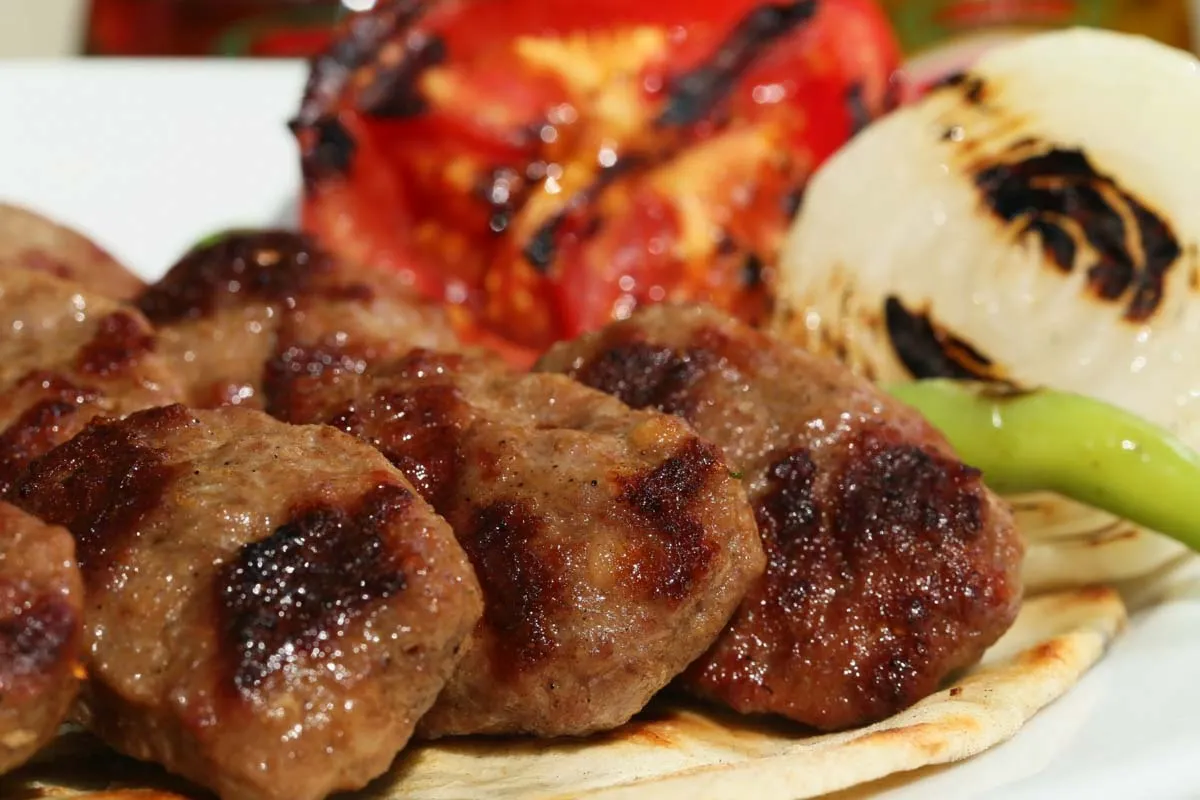 Close-up of grilled köfte on flatbread with charred tomato, grilled onion, and a green pepper