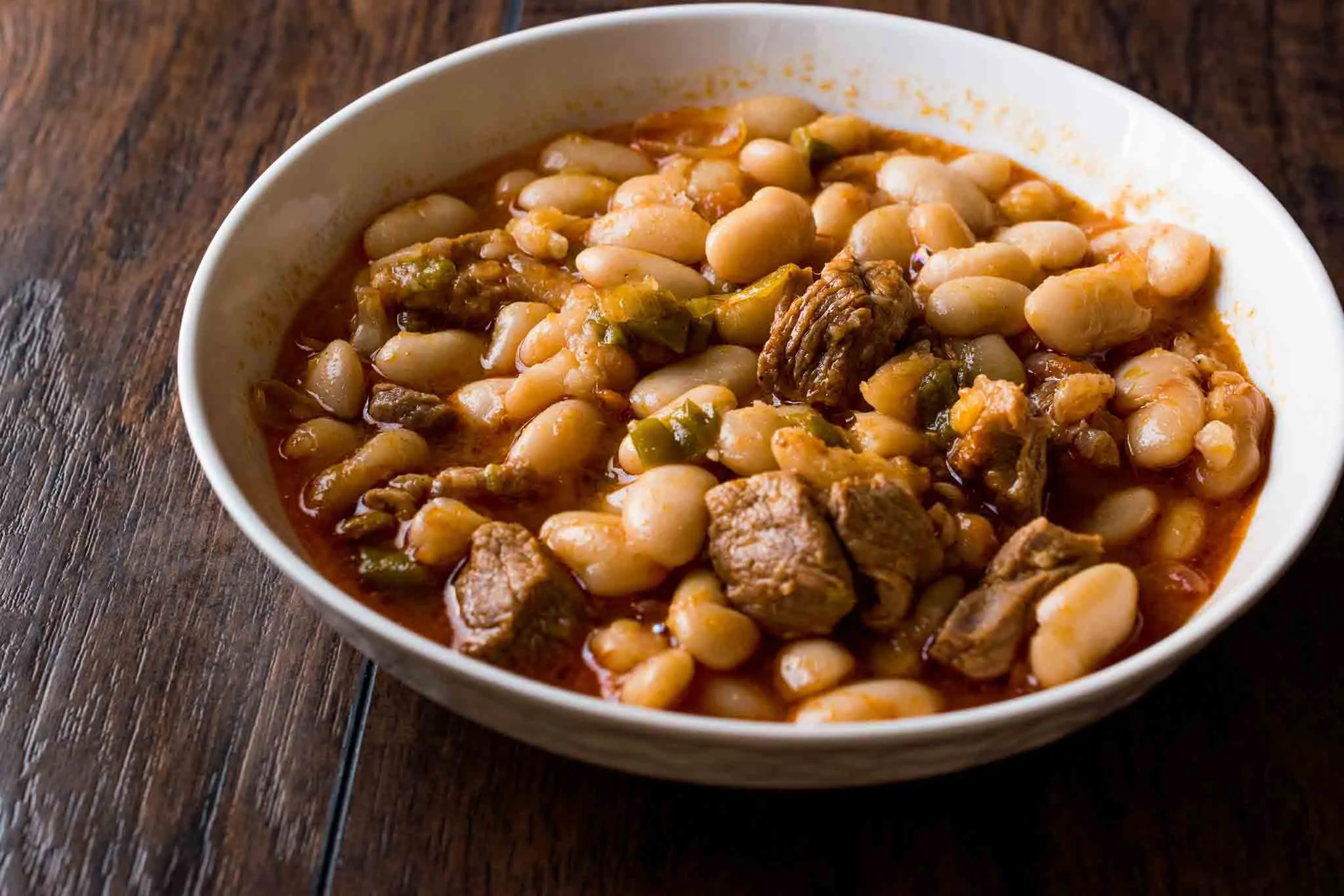 White bowl of Turkish white bean stew with lamb chunks and green peppers in tomato sauce on a wooden table