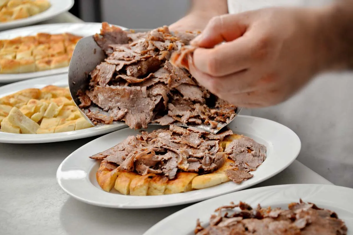 A cook layers thin sliced doner meat over pieces of pide bread on white plates