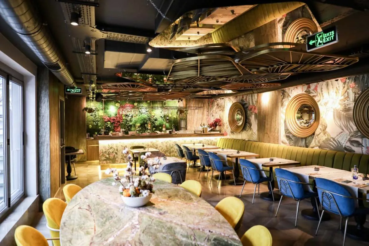Restaurant interior with green velvet booths, blue and yellow chairs, marble tables, and a plant wall