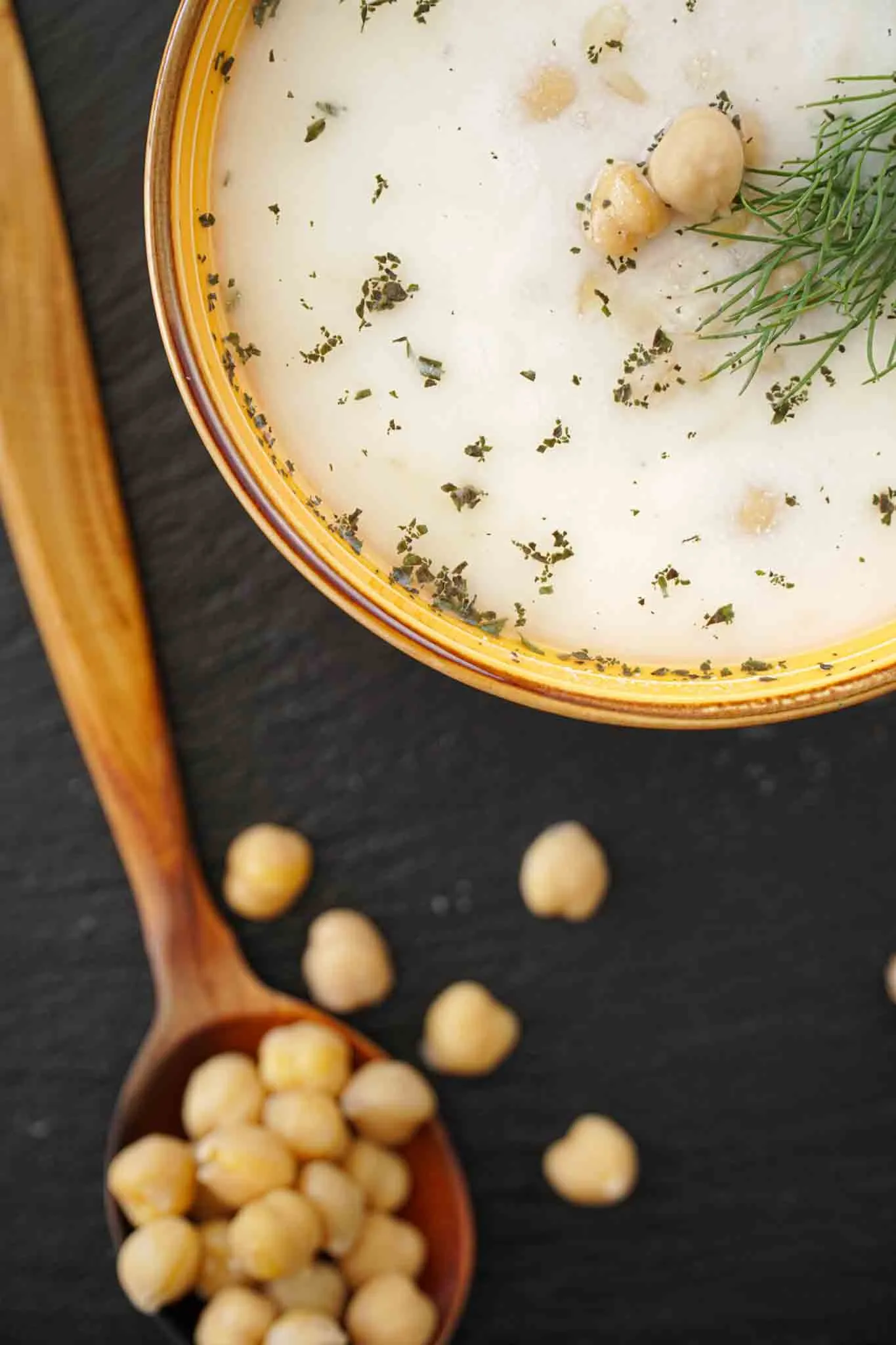 Creamy white yogurt soup with chickpeas and dill in a yellow bowl beside a wooden spoon
