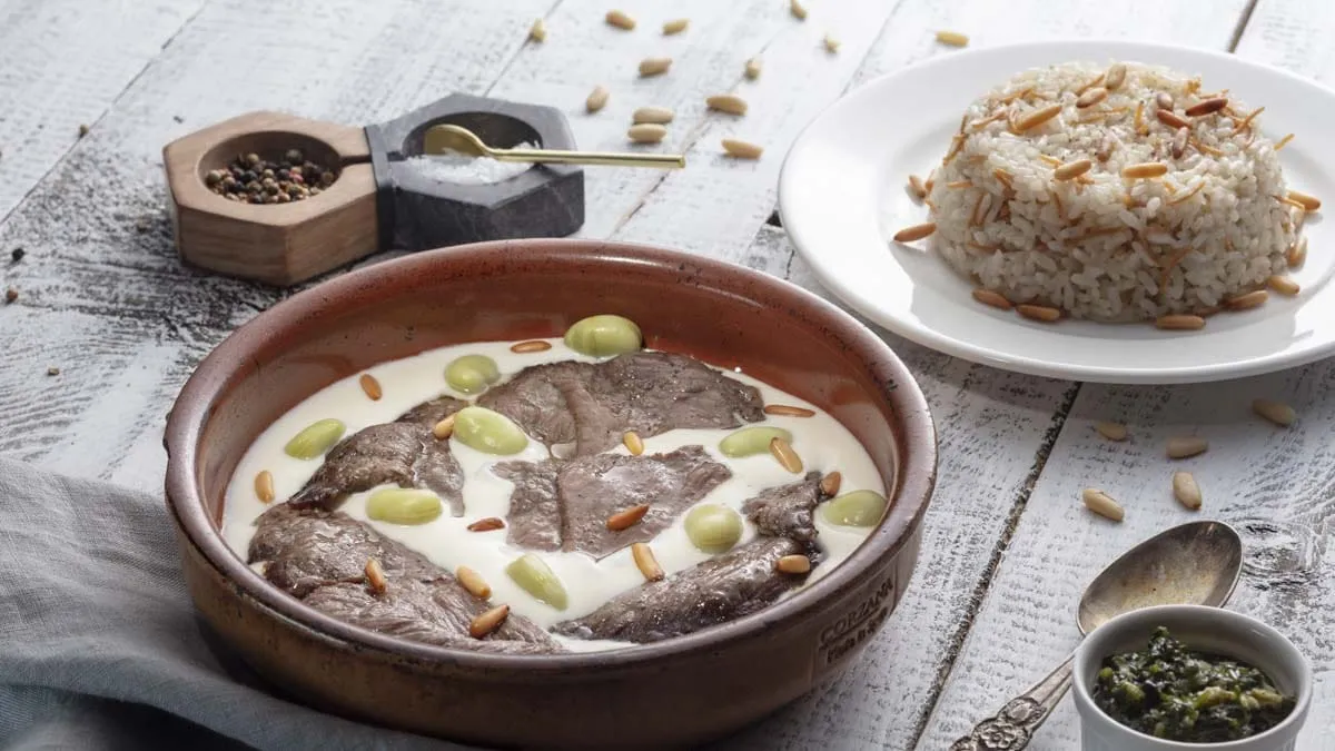 Sliced lamb in yogurt sauce with pine nuts beside a plate of rice pilaf