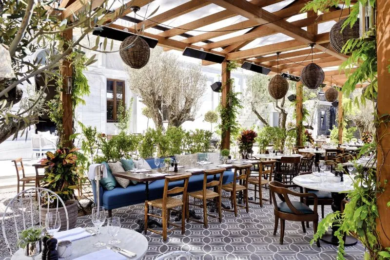 Outdoor restaurant terrace with a wooden pergola, woven lantern shades, olive trees, and set tables on patterned tile