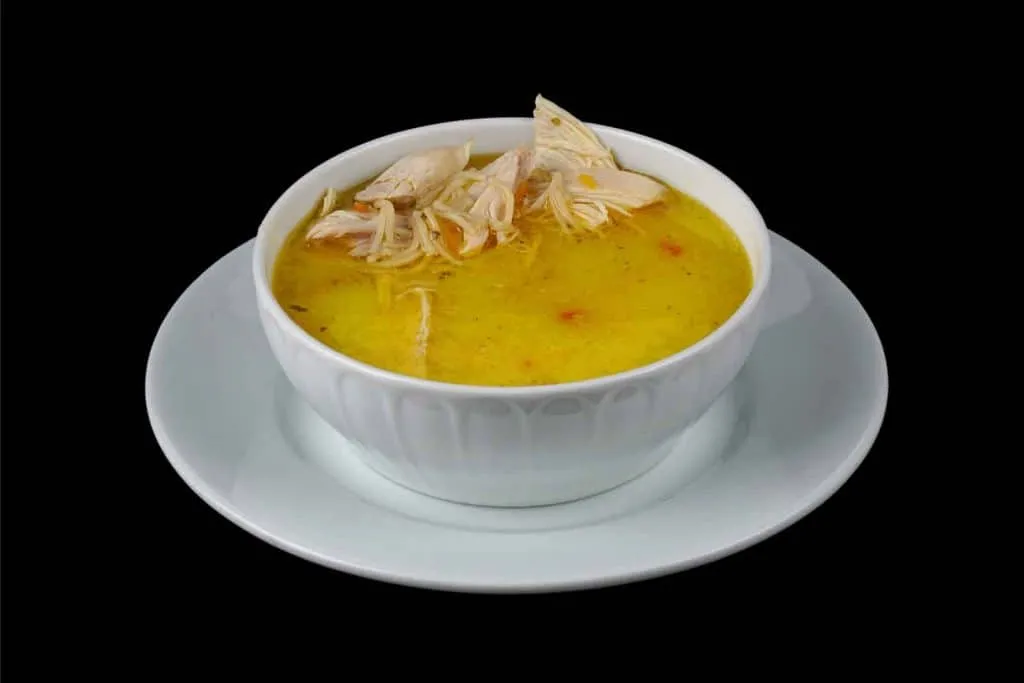 White bowl of golden chicken soup topped with shredded chicken on a saucer
