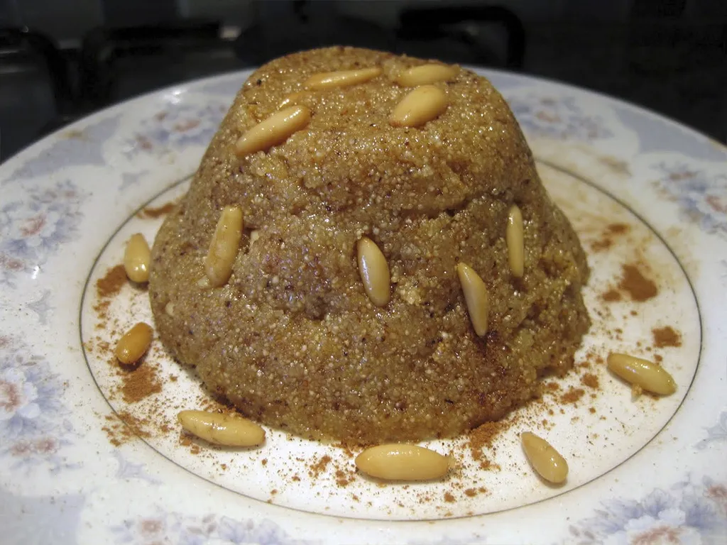 Conical portion of irmik helvasi (semolina dessert) on blue plate sprinkled with cinnamon and pine nuts