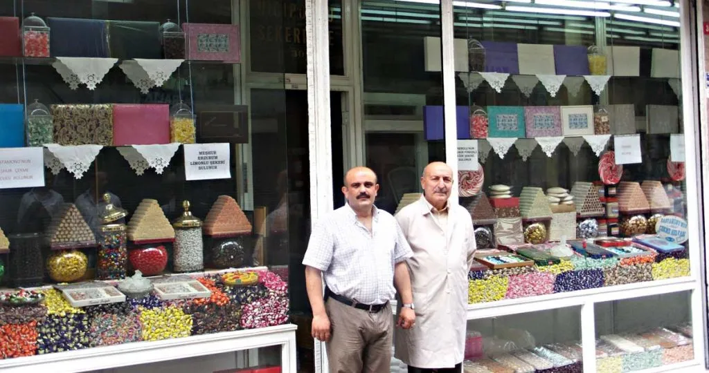 Two shopkeepers outside Hicipoğlu Şekerleme, its windows stacked with wrapped candies and helva