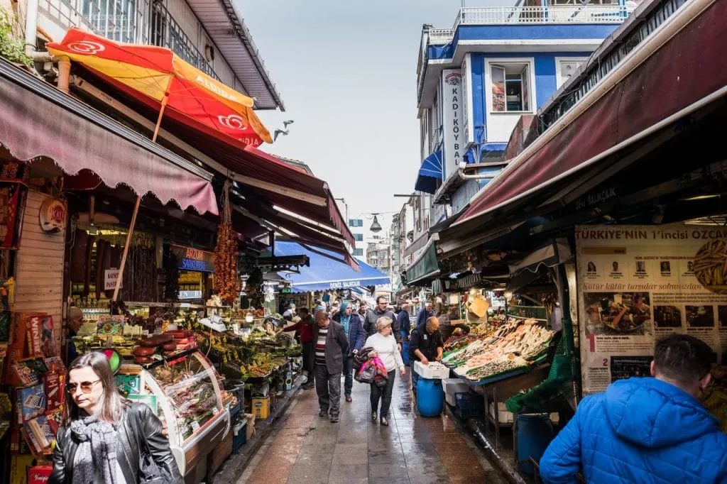 Shoppers wandering busy kadikoy market street looking at fruit shop and fresh fish vendor, Istanbul tour