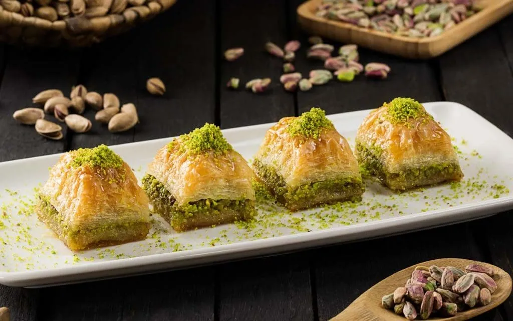 Four pieces of glossy pistachio baklava on a white plate with loose pistachios scattered around
