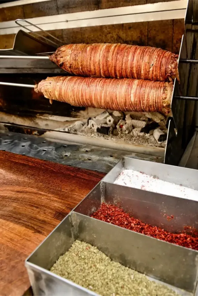Two kokoreç rolls roasting on spits at Kral Kokoreç, with trays of salt, chili flakes, and oregano below