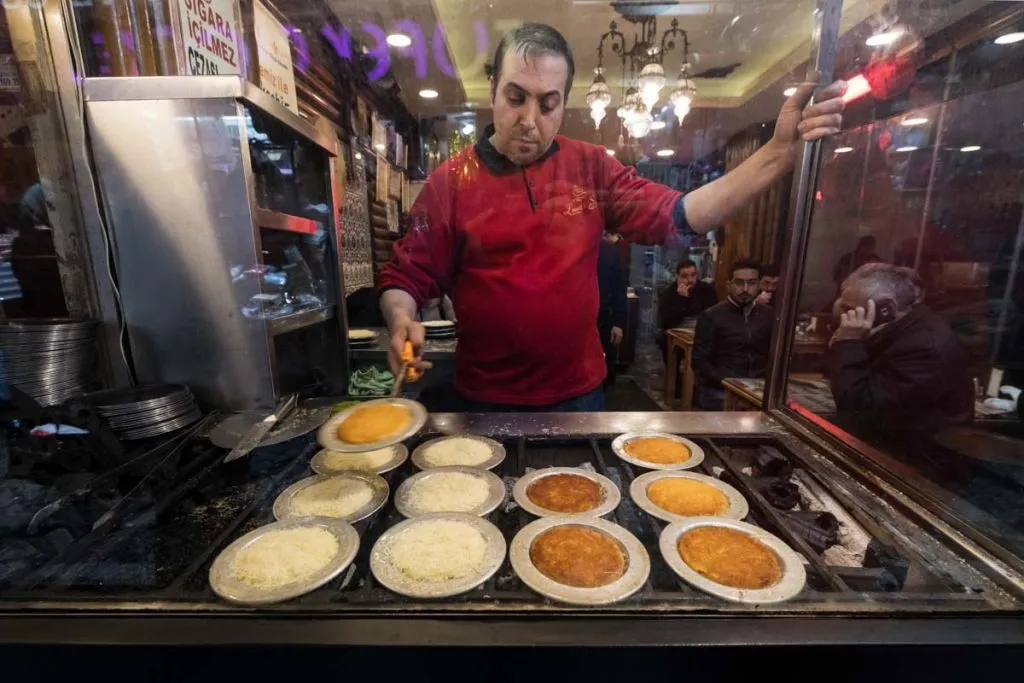 Cook turning künefe trays on a window griddle, rows of künefe baking from pale to deep golden