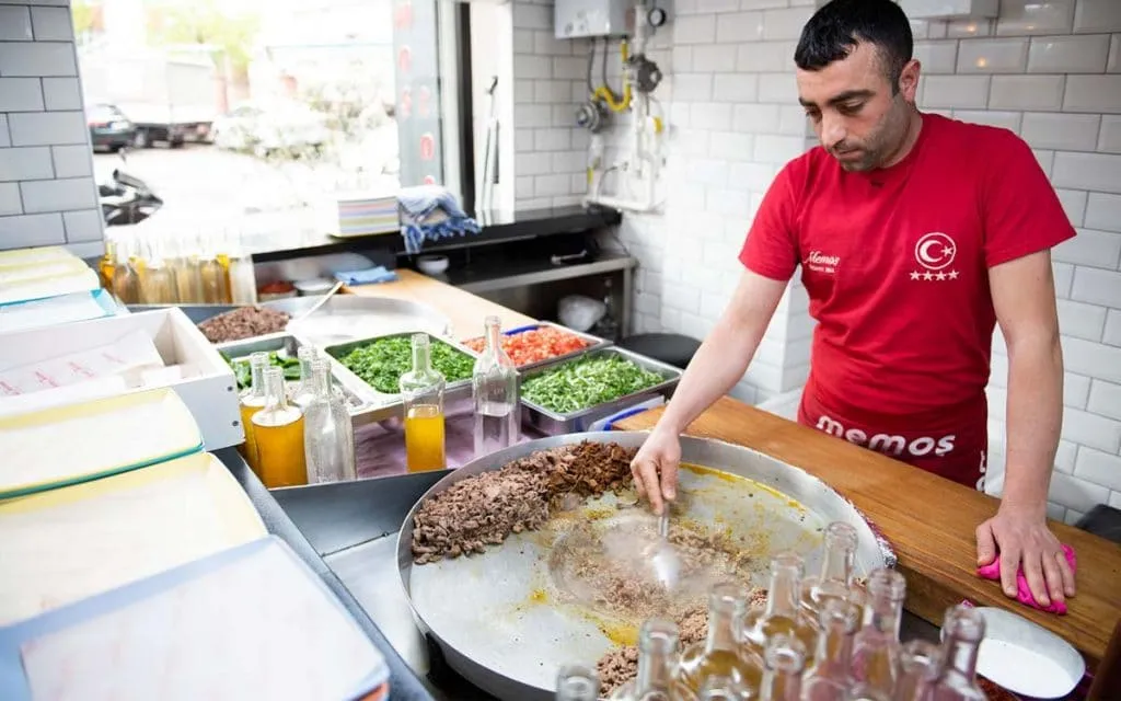 Cook in a red Memoş shirt working chopped beef on a round sac griddle, with trays of parsley, tomato, and onion behind the counter