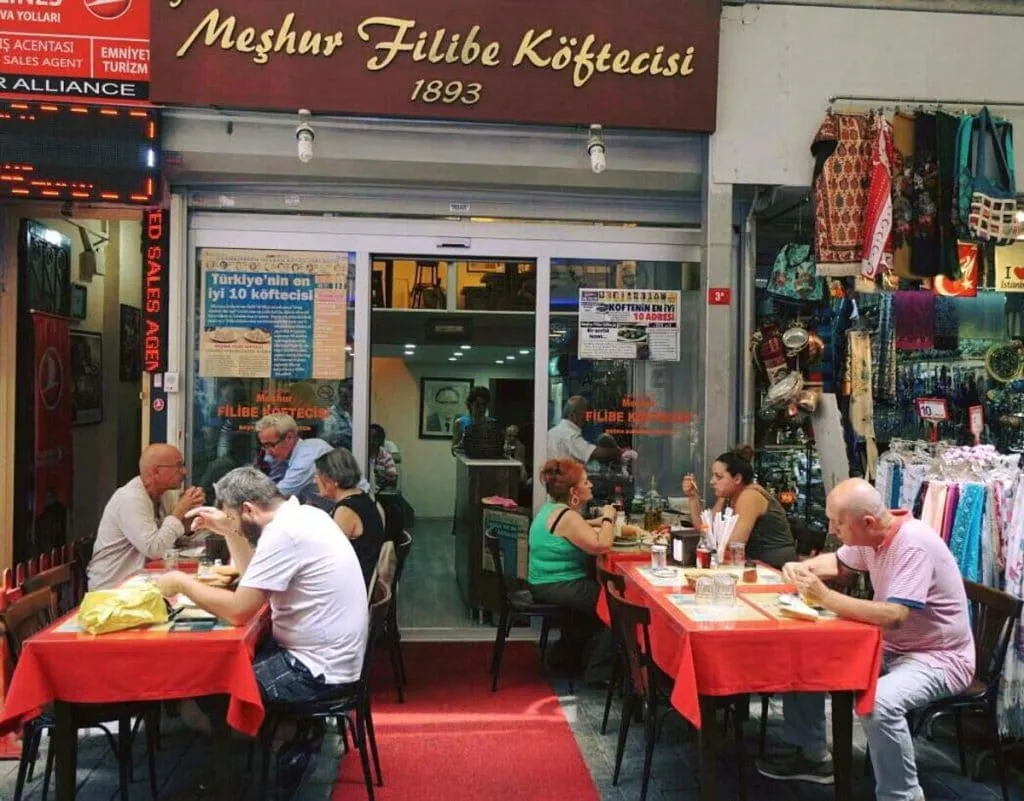 Diners at red-clothed sidewalk tables outside Meşhur Filibe Köftecisi, under its gold 1893 storefront sign