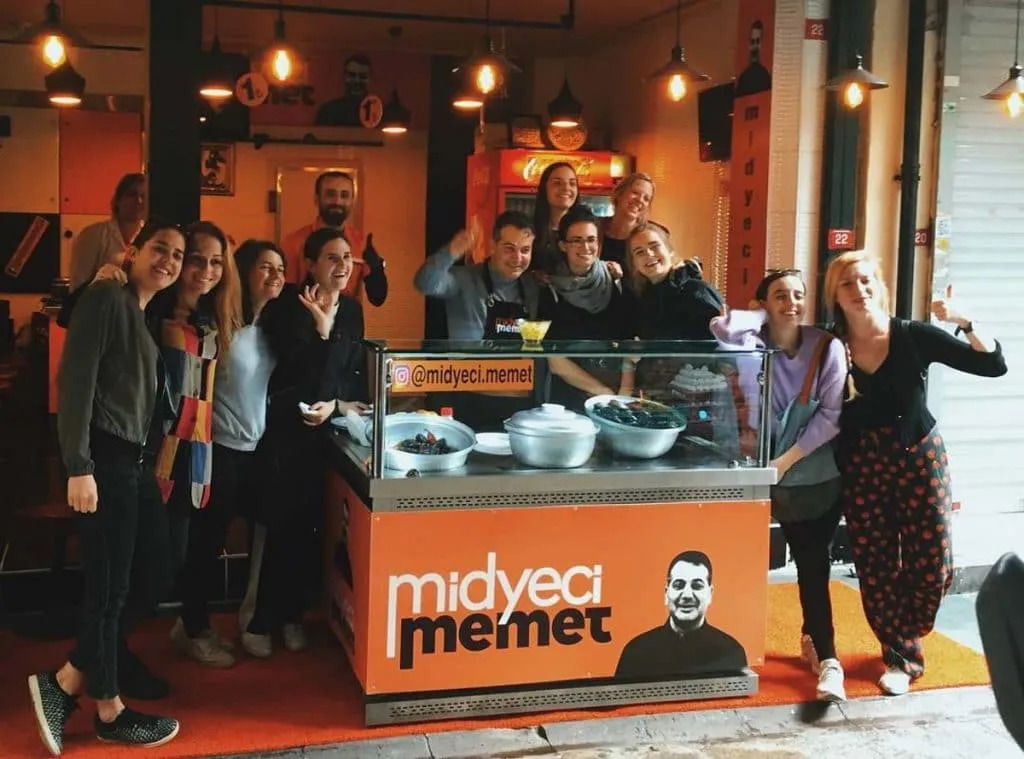 A group of guests gathered around the orange Midyeci Memet mussel cart in front of the shop