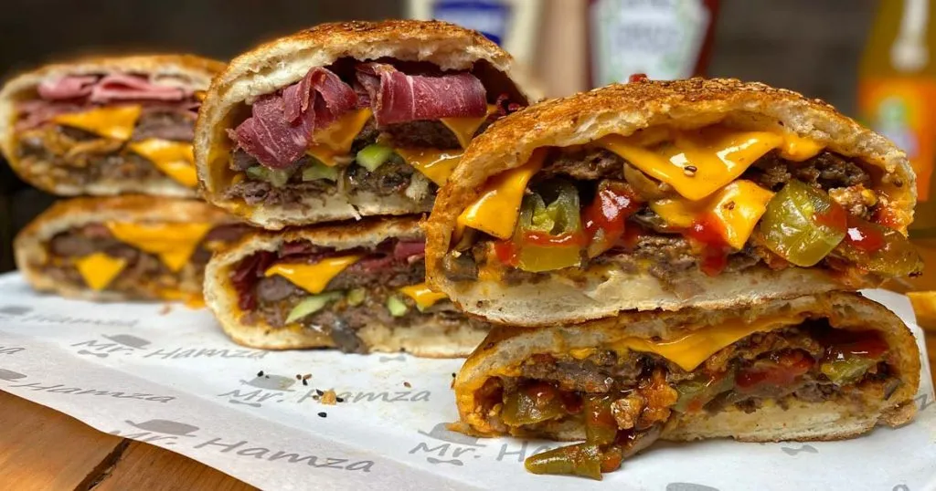 Halved HAMZA pizza-burgers stacked on Mr. Hamza branded paper, showing layers of beef, melted cheddar, pickles, and pastrami inside the dough