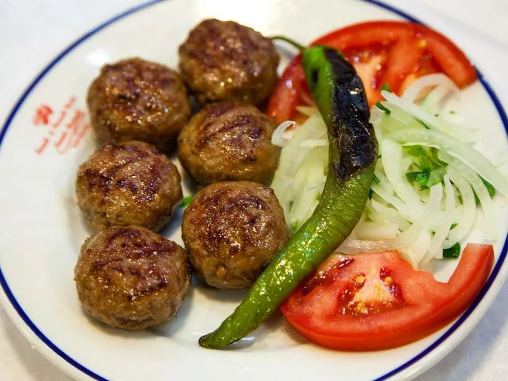 Six grilled köfte on a plate at Namlı Rumeli Köftecisi with tomato slices, shaved onion, and a charred green pepper
