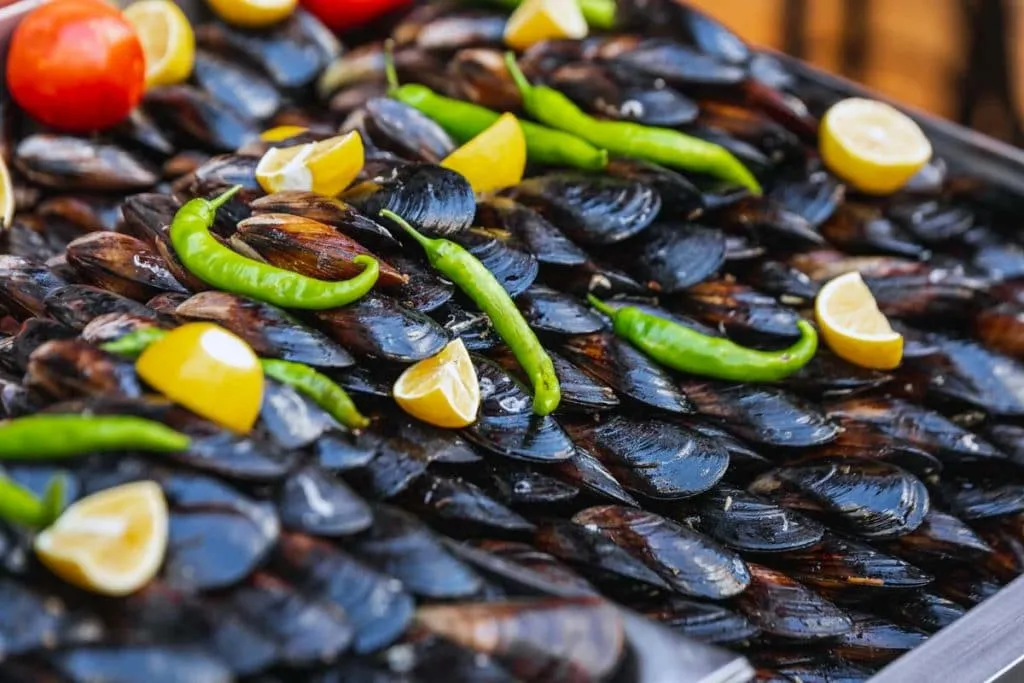 Rows of stuffed mussels stacked on a street cart tray, topped with lemon wedges and green peppers