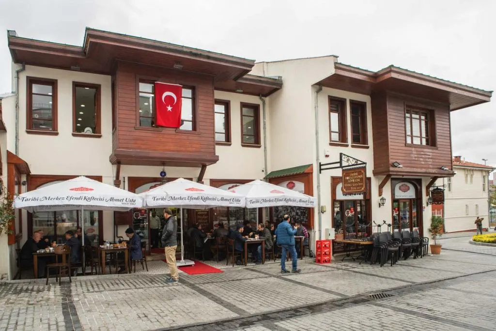 Wooden two-story building of Tarihi Merkez Efendi Köftecisi with branded white umbrellas over its sidewalk tables
