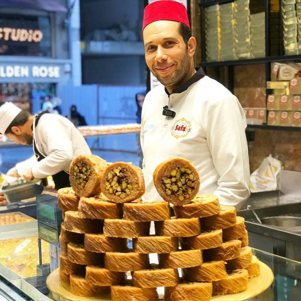 Smiling server in a red fez at Tatlıcı Safa behind a stacked pyramid of pistachio-filled kadayıf rings