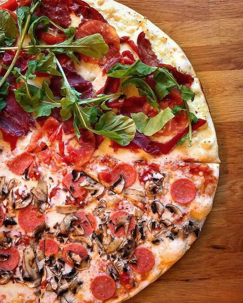 Overhead view of a half-and-half thin-crust pizza at The Upper Crust, cured beef and arugula on one side, pepperoni and mushrooms on the other