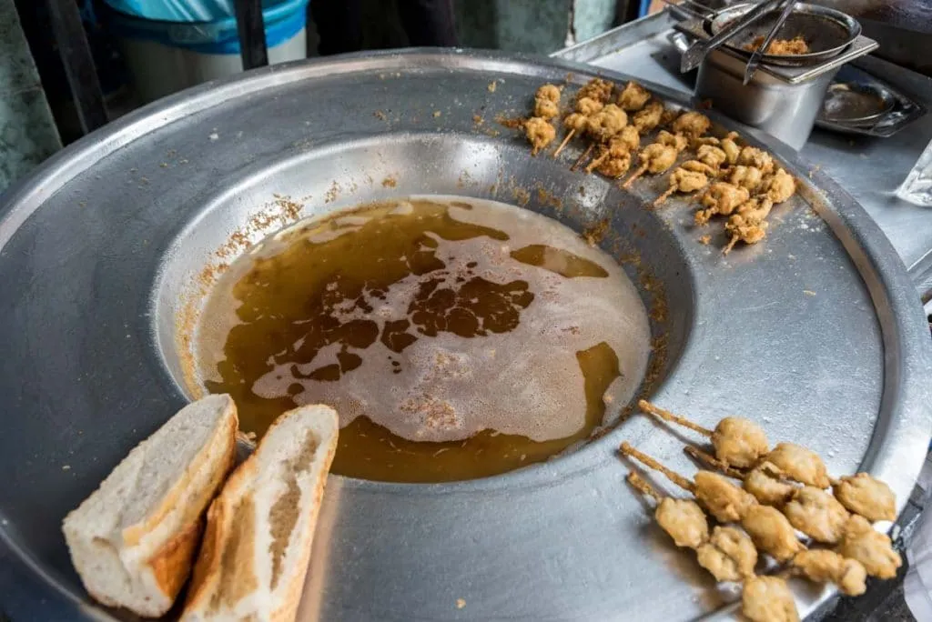 Skewers of fried mussels resting on the rim of a street fryer next to halved loaves of bread