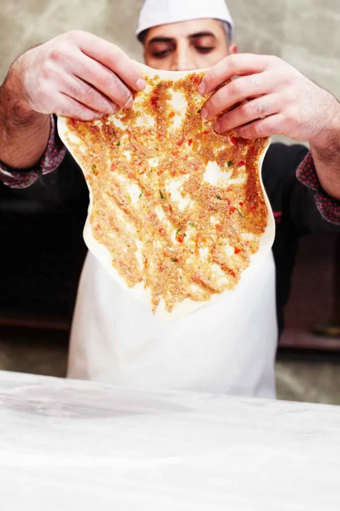 Usta holding up a thin round of raw lahmacun dough topped with hand-minced meat before baking
