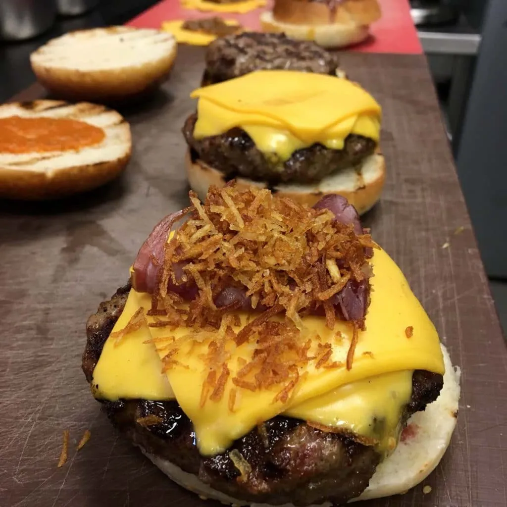 Zapata Burger cheeseburgers mid-assembly, the nearest piled with crispy shoestring potatoes and red onion over cheddar